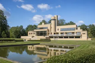 Villa Cavrois modernist building with yellow brick facade, large windows, surrounded by green lawns and hedges, under a blue sky with soft daylight.
