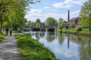 Canal de Roubaix with a tree-lined towpath, a family walking, two cyclists, calm water, and old brick buildings under a blue spring sky.