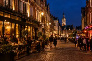 Central street in Lille at night with brick buildings lit by warm streetlights, people enjoying terraces and walking, relaxed nightlife atmosphere.